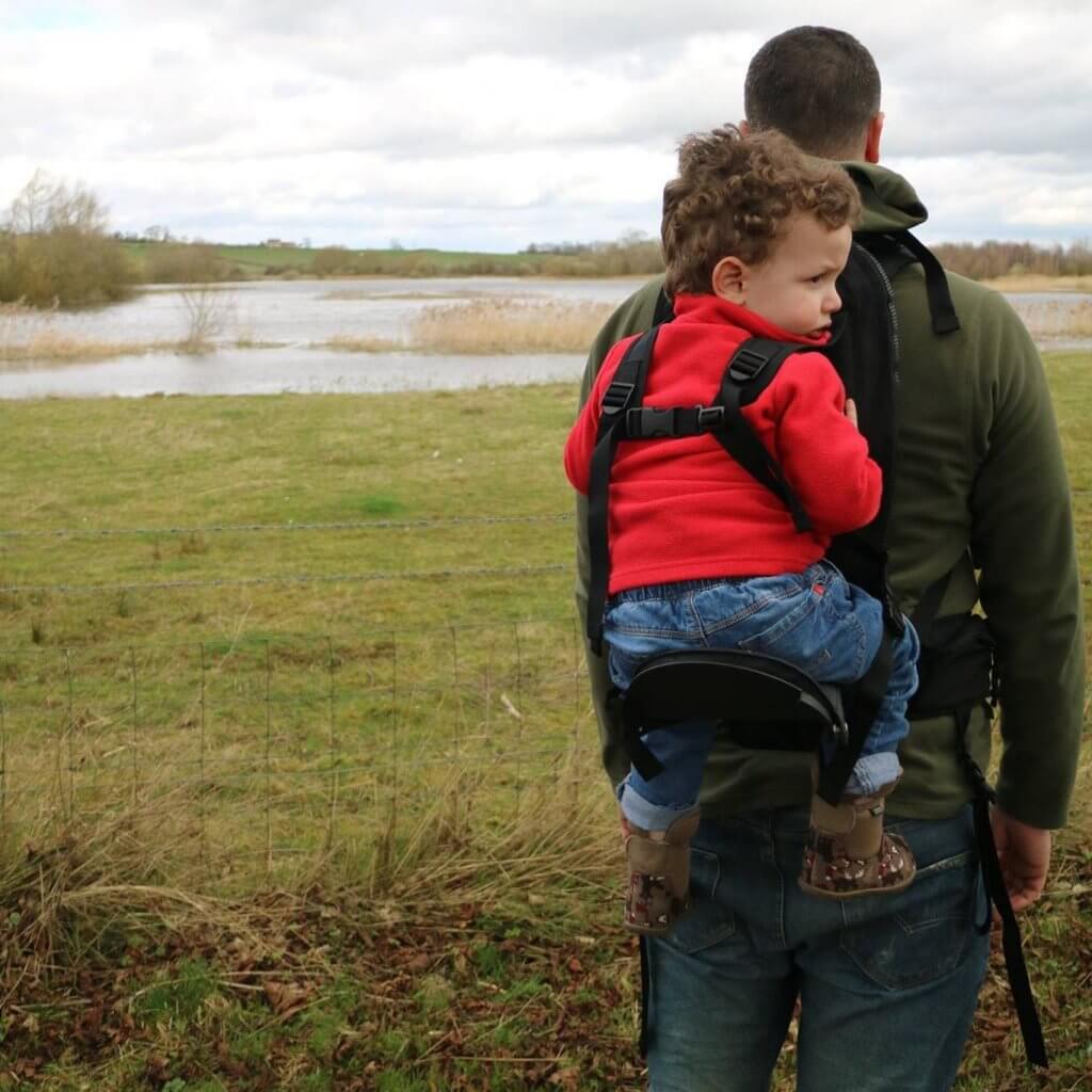 A man looks out over a lake, a toddler is in a carrier on his back