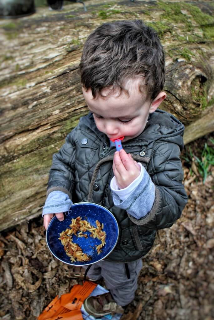A young boy eats some pasta from a bowl. He is outdoors, standing by a fallen log